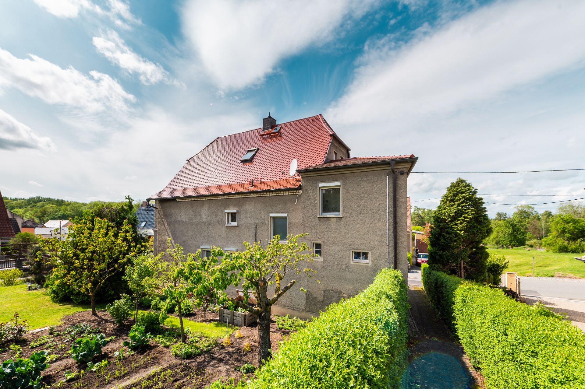 Zweistöckiges Wohnhaus mit rotem Dach, Garten und Hecke im Vordergrund bei sonnigem Wetter.