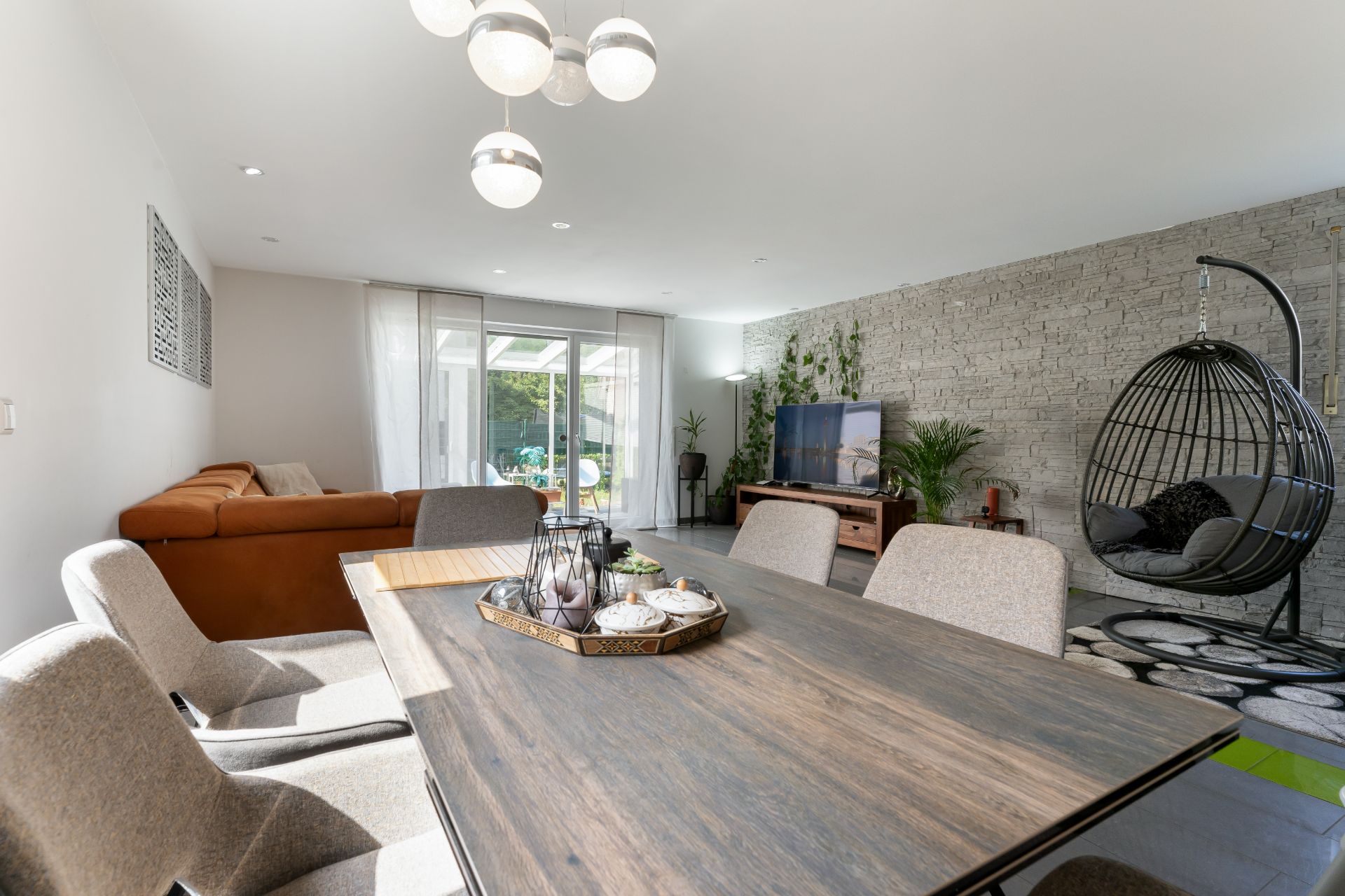 Helles Wohnzimmer mit Esstisch, grauen Stühlen, Sofa, Fernseher und Hängesessel.