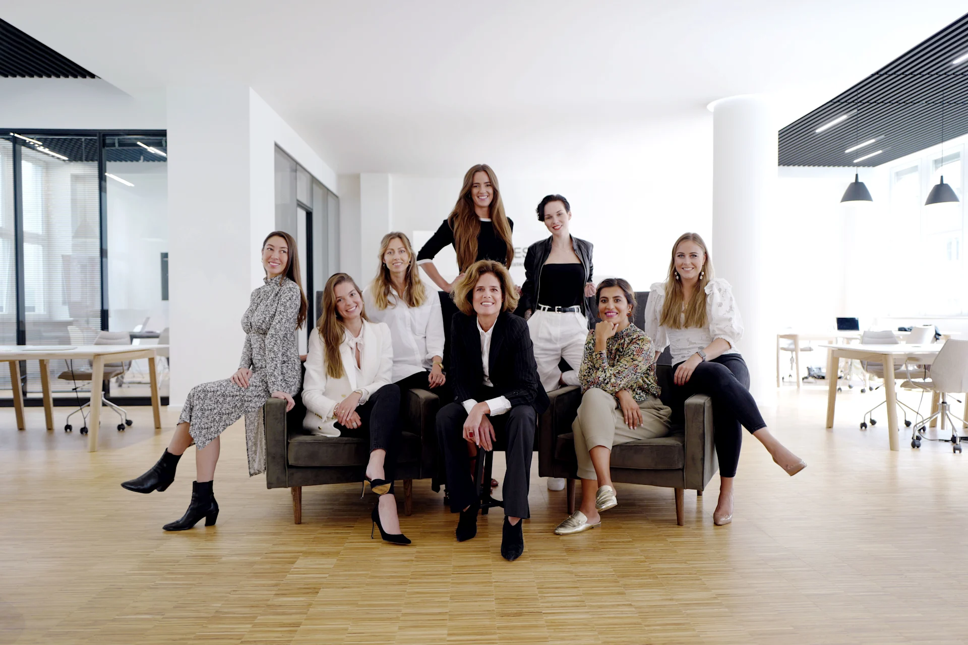 Neun Frauen sitzen und stehen in einem modernen, hellen Büroraum mit Holzboden.