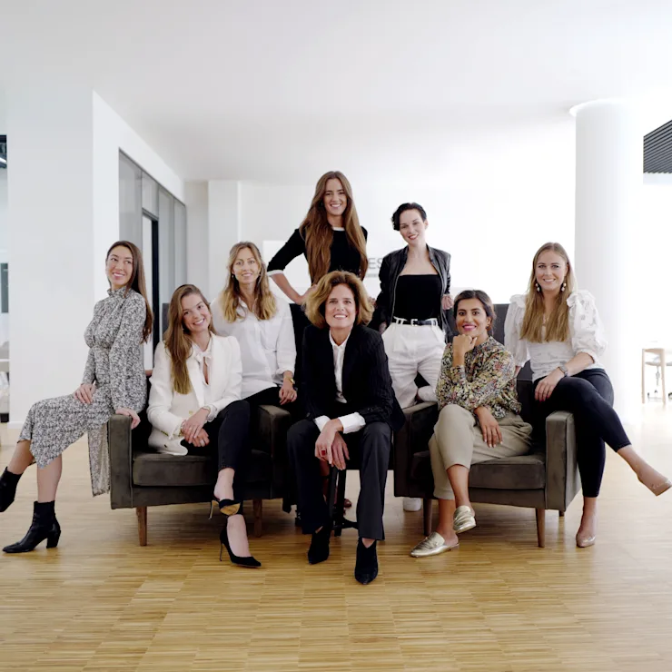 Neun Frauen sitzen und stehen in einem modernen, hellen Büroraum mit Holzboden.