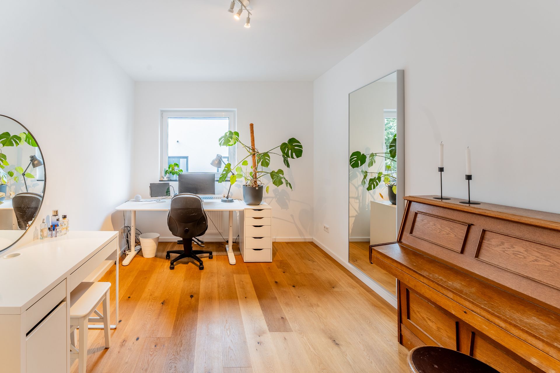 Helles Zimmer mit Schreibtisch, Stuhl, Klavier, großem Spiegel und Zimmerpflanzen auf Holzboden.