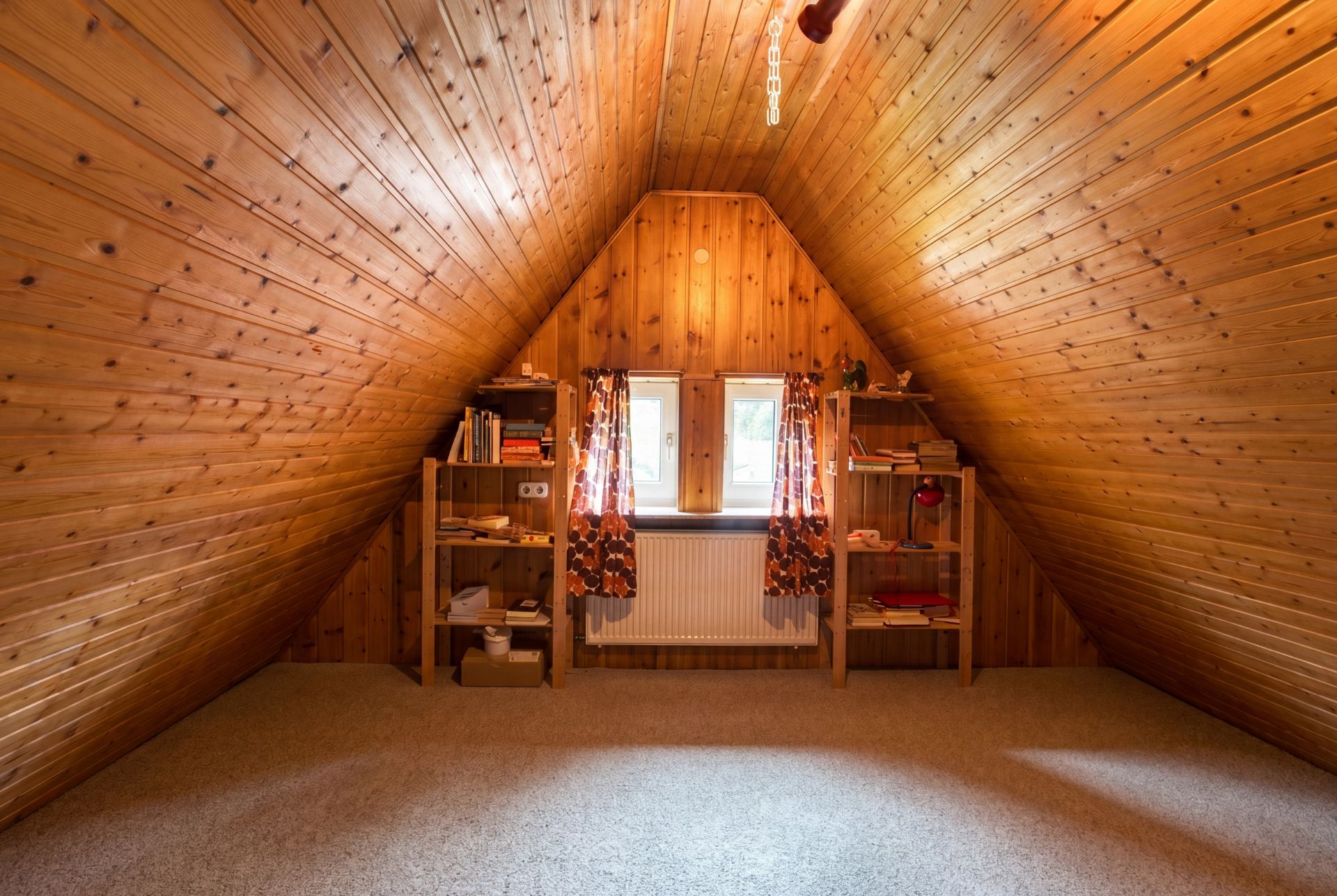 Dachzimmer mit Holzverkleidung, zwei Fenstern, Vorhängen und zwei Bücherregalen.