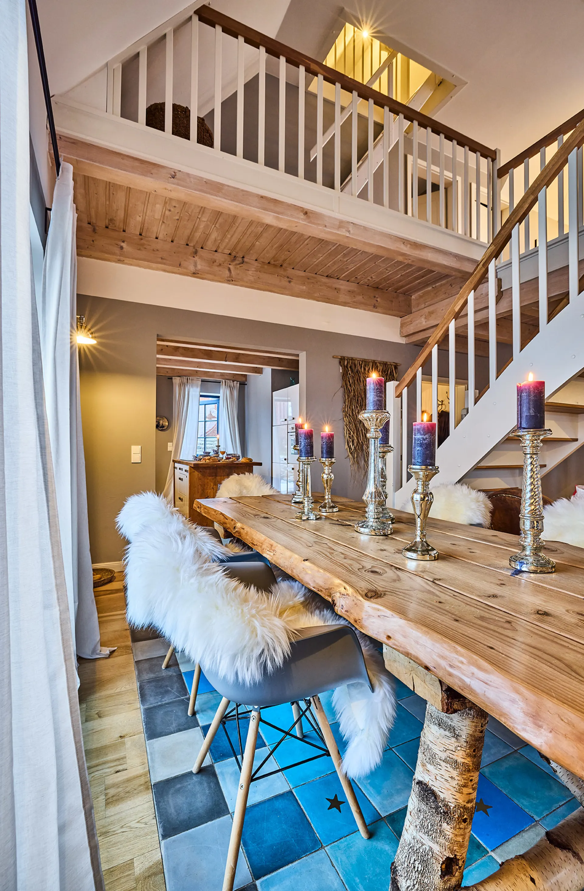 Essbereich mit Holztisch, Kerzen und Stühlen mit Fell, Treppe und Galerie im Hintergrund.