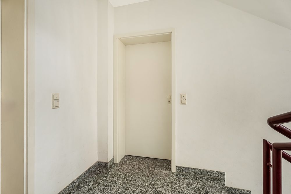 Heller Flur mit weißer Tür, Lichtschaltern und Treppe mit rotem Geländer.