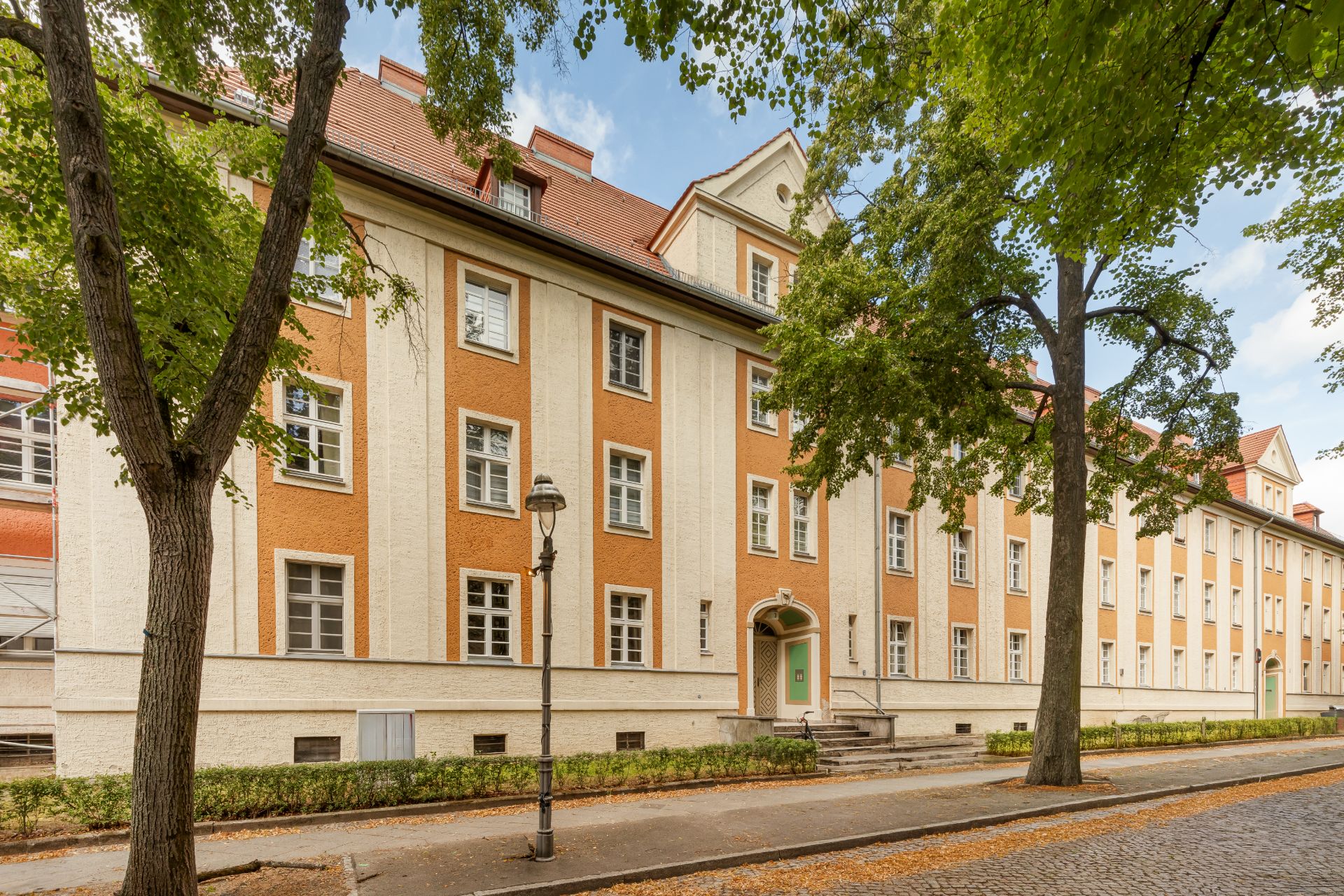 Mehrstöckiges Wohngebäude mit orange-weißer Fassade und Eingangsbereich, Bäume an der Straße.