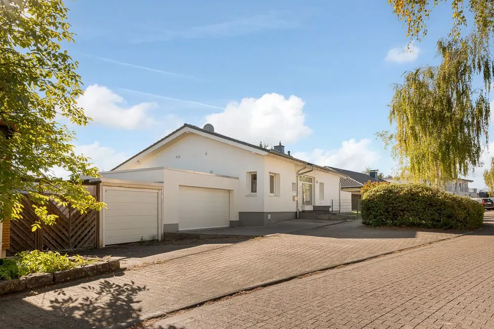 Weißes Einfamilienhaus mit Garage und Einfahrt, umgeben von Bäumen und Sträuchern.