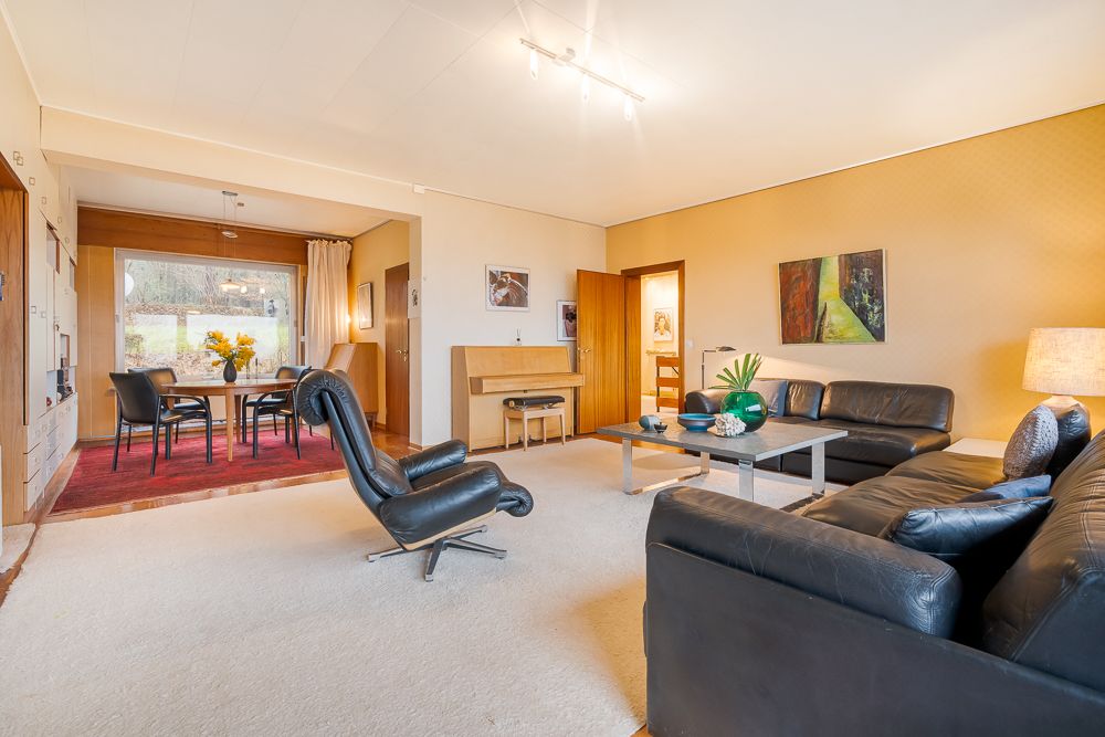 Helles Wohnzimmer mit schwarzen Ledersofas, Sessel, Couchtisch und Essbereich mit großem Fenster.