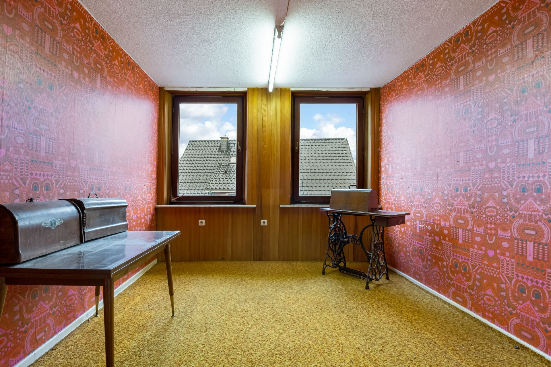 Raum mit orangener Tapete, zwei Fenstern, vintage Nähmaschinen und gelbem Teppichboden.
