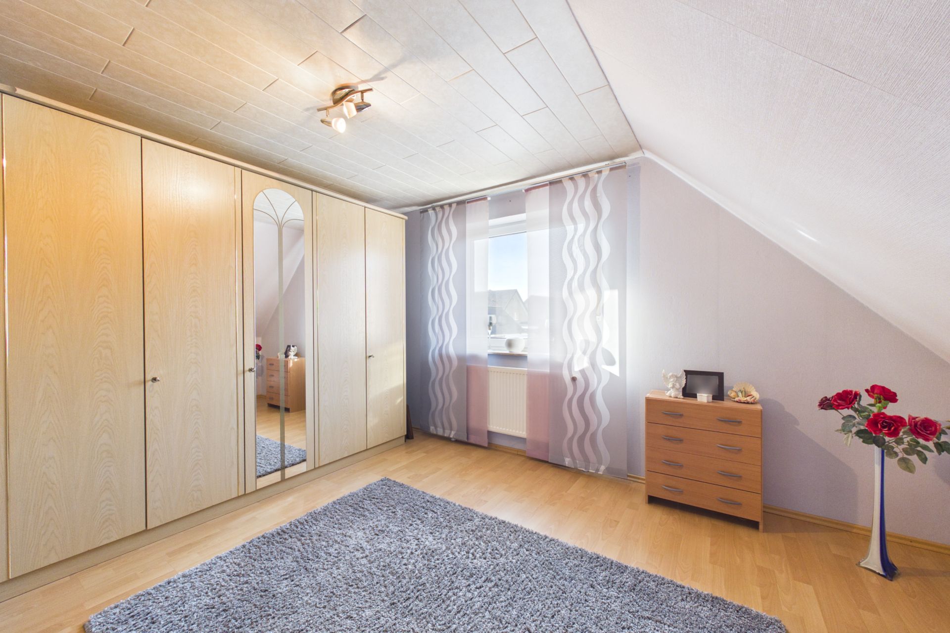 Helles Schlafzimmer mit Kleiderschrank, Kommode, Teppich und Fenster mit Vorhängen.