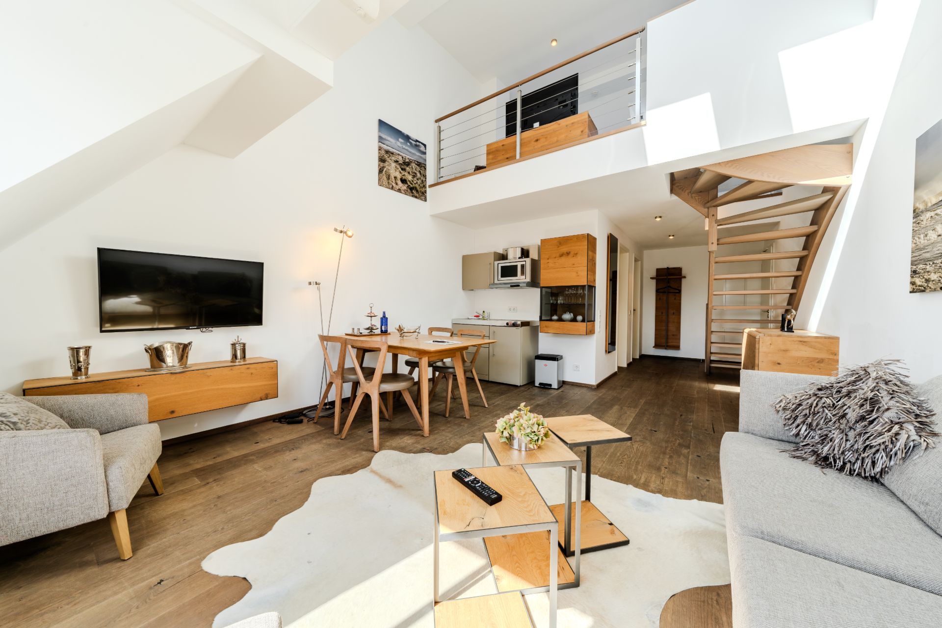 Helles Wohnzimmer mit Sofa, Esstisch, Fernseher, Küche und Treppe zu einer offenen Galerie.