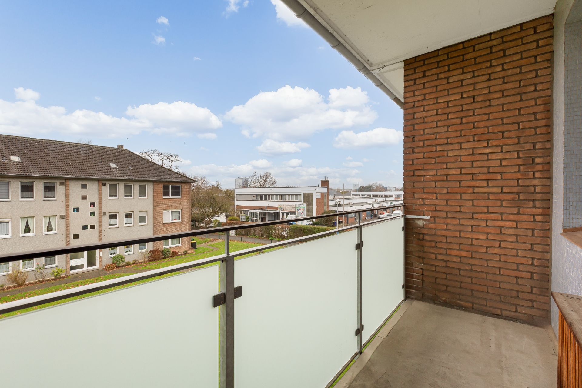 Balkon mit Glasgeländer, Blick auf Wohnhäuser und grüner Außenbereich.