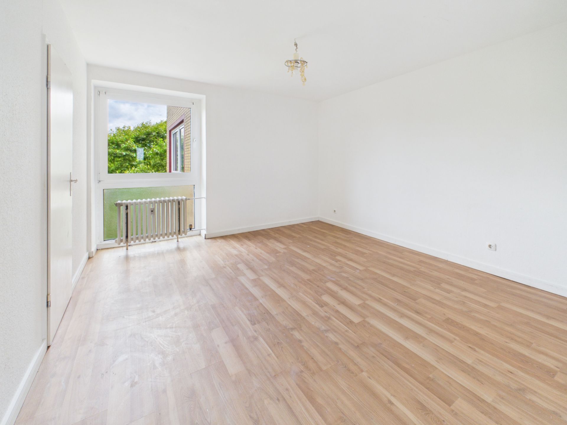 Heller, leerer Raum mit Holzfußboden, weißer Wand und großem Fenster mit Blick ins Grüne.