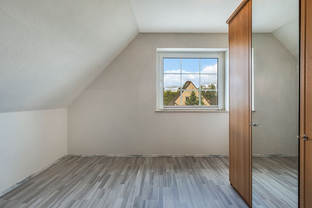Helles Zimmer mit Dachschräge, Fenster und einem Kleiderschrank mit Spiegeltür.