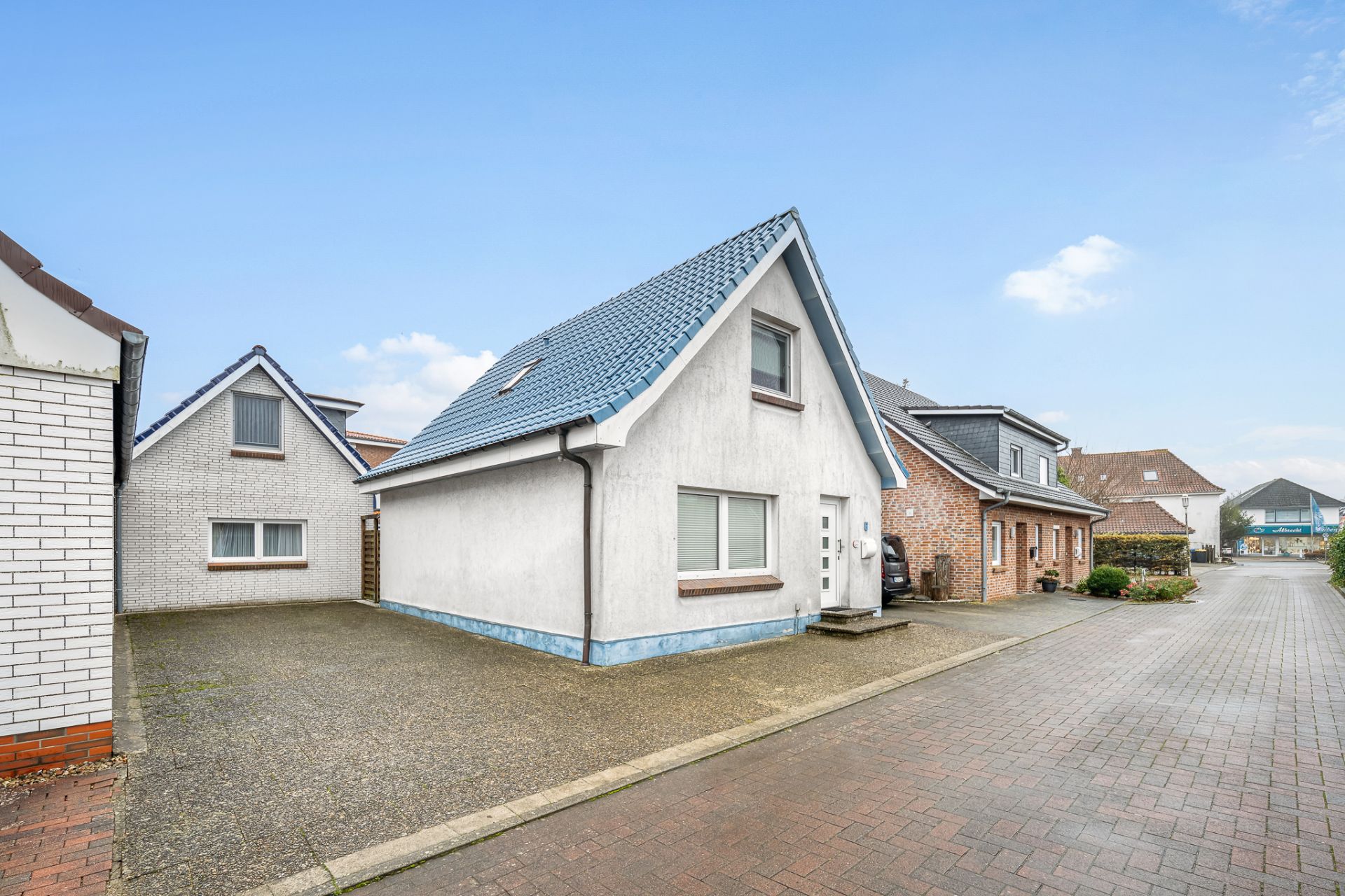 Weißes Einfamilienhaus mit blauem Dach und gepflastertem Hof in einer Wohnsiedlung.