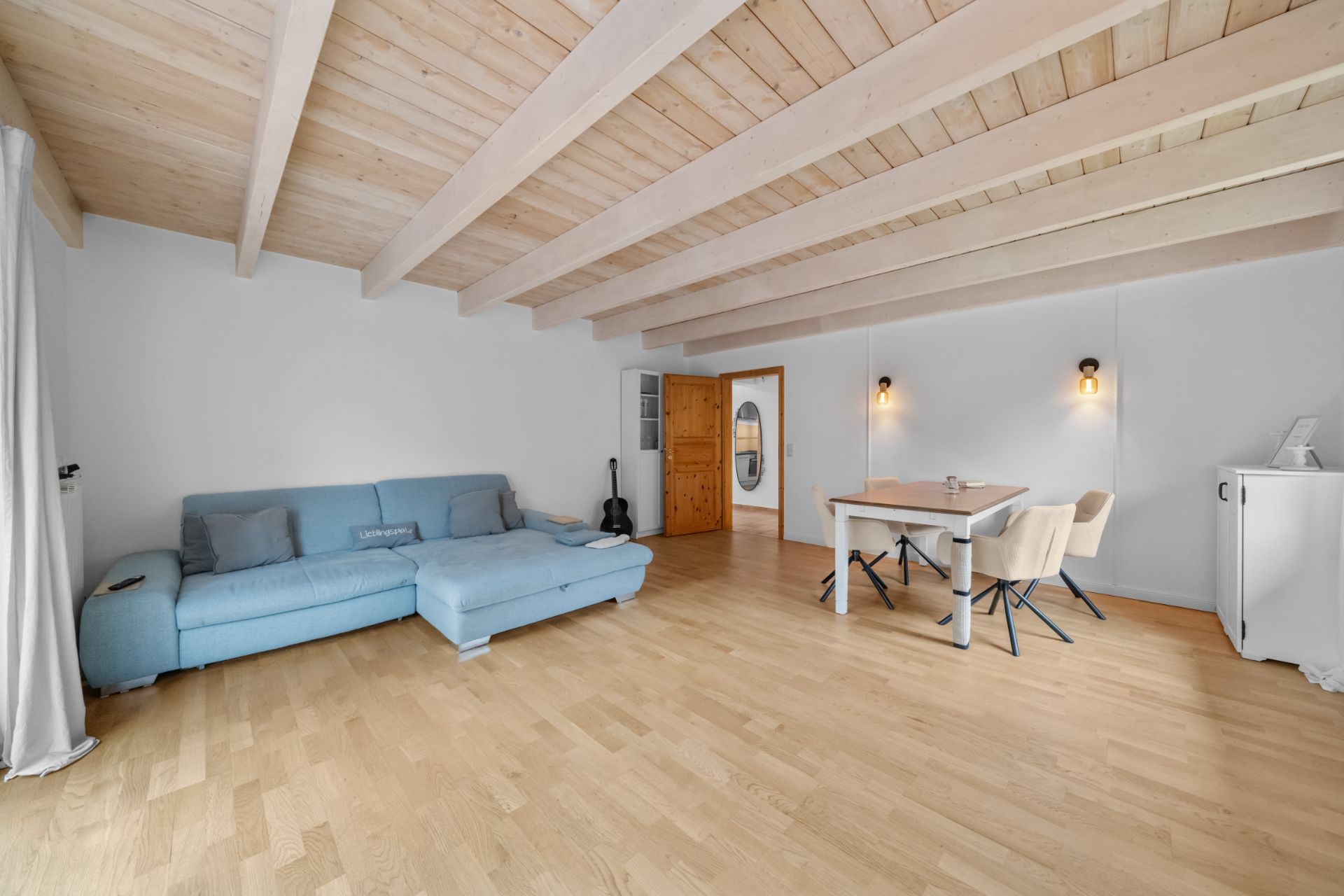 Wohnzimmer mit hellblauem Sofa, Esstisch mit vier Stühlen, Holzboden und sichtbaren Deckenbalken.