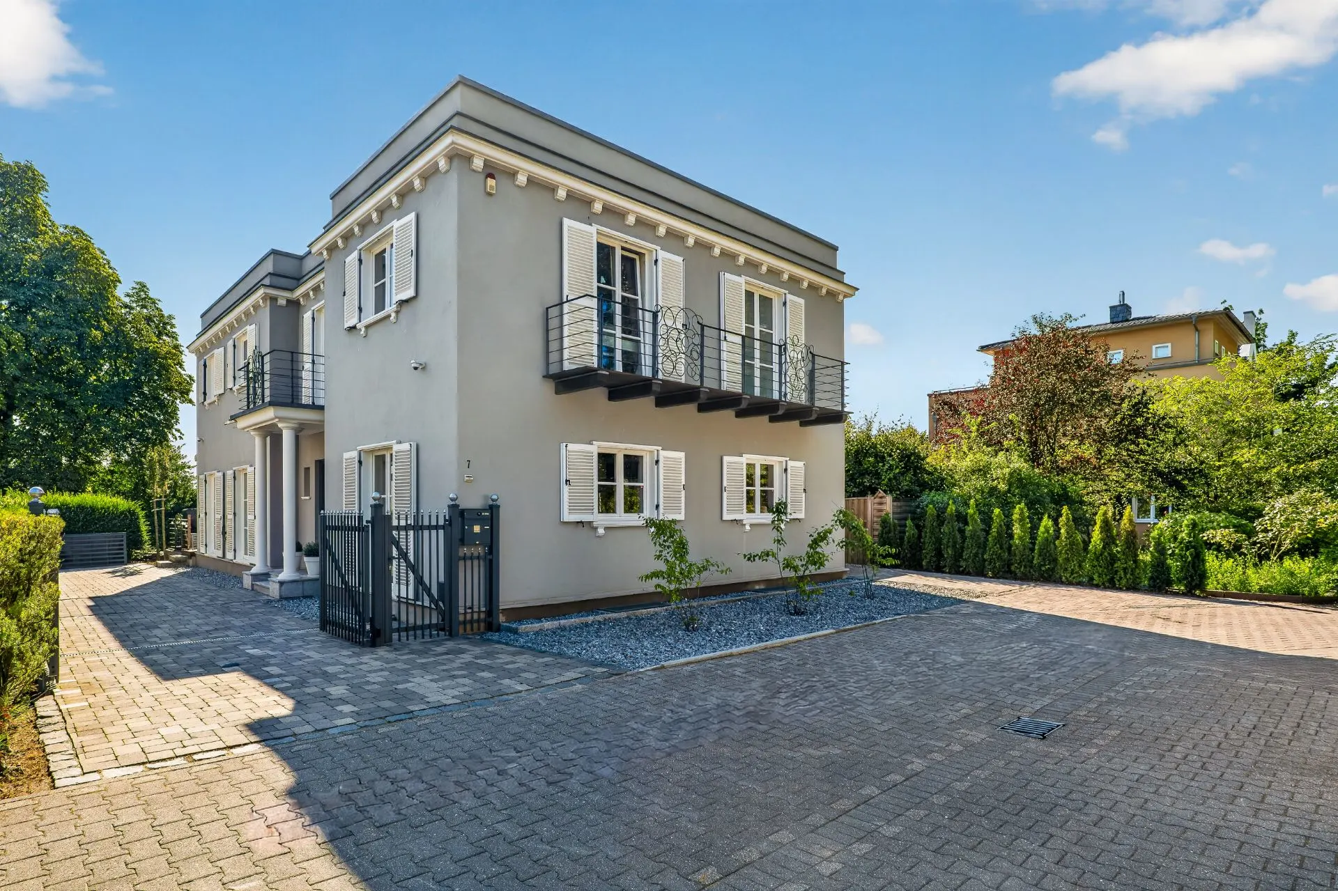 Zweistöckiges Wohnhaus mit Balkonen und weißen Fensterläden, umgeben von Hof und Garten.