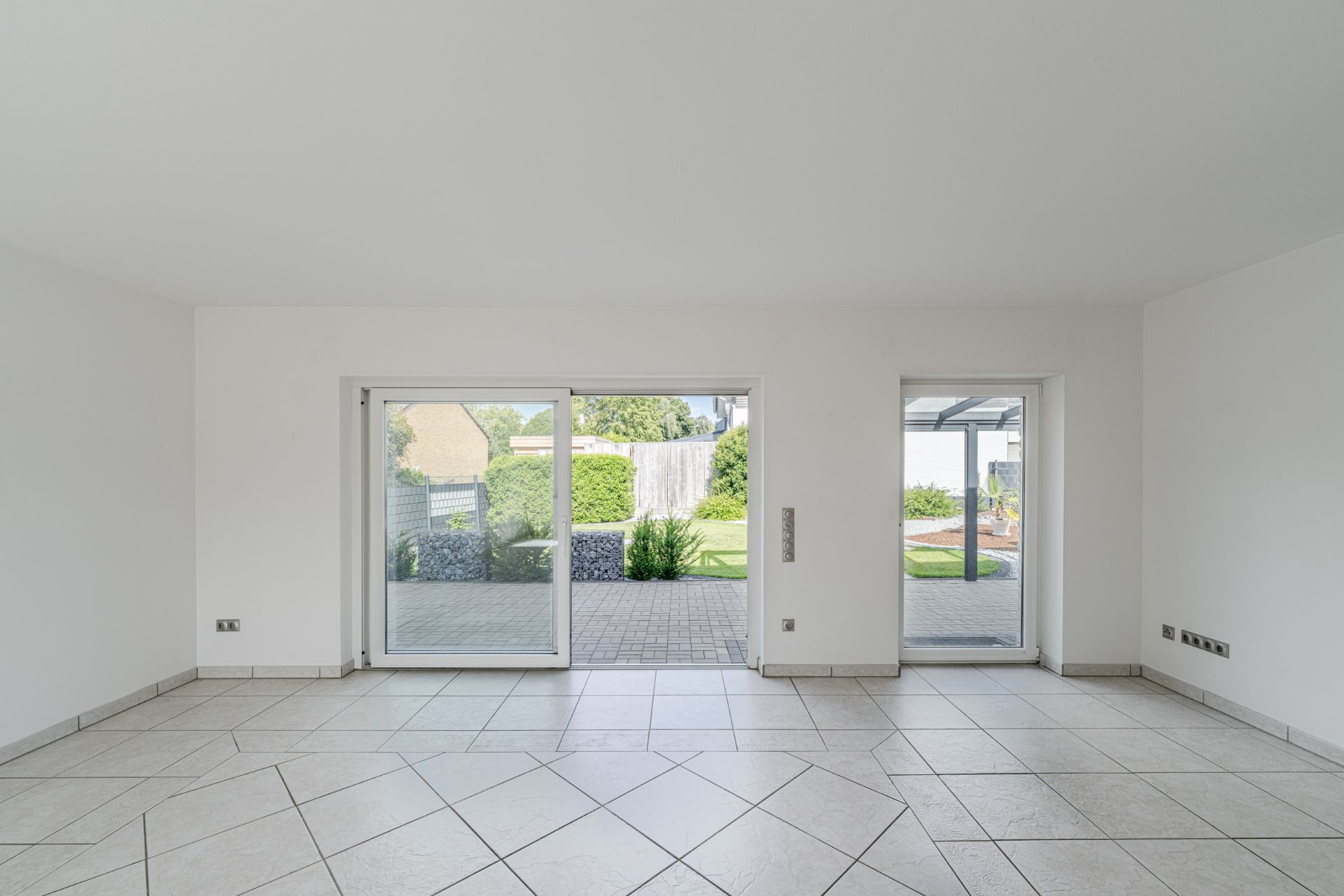 Helles Wohnzimmer mit großen Fliesen, weißen Wänden und bodentiefen Fenstern zum Garten.