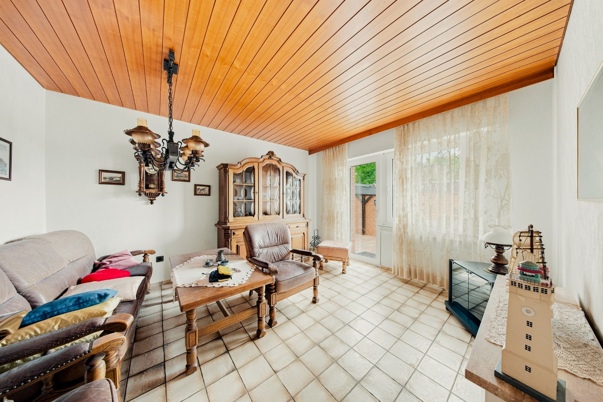 Helles Wohnzimmer mit Holzmöbeln, Sofas, Vitrine und großen Fenstern mit Vorhängen.