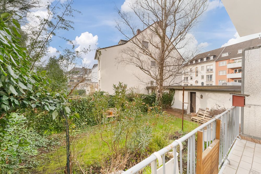 Blick von einem Balkon auf einen grünen Garten mit Sträuchern, Bank und angrenzenden Häusern.