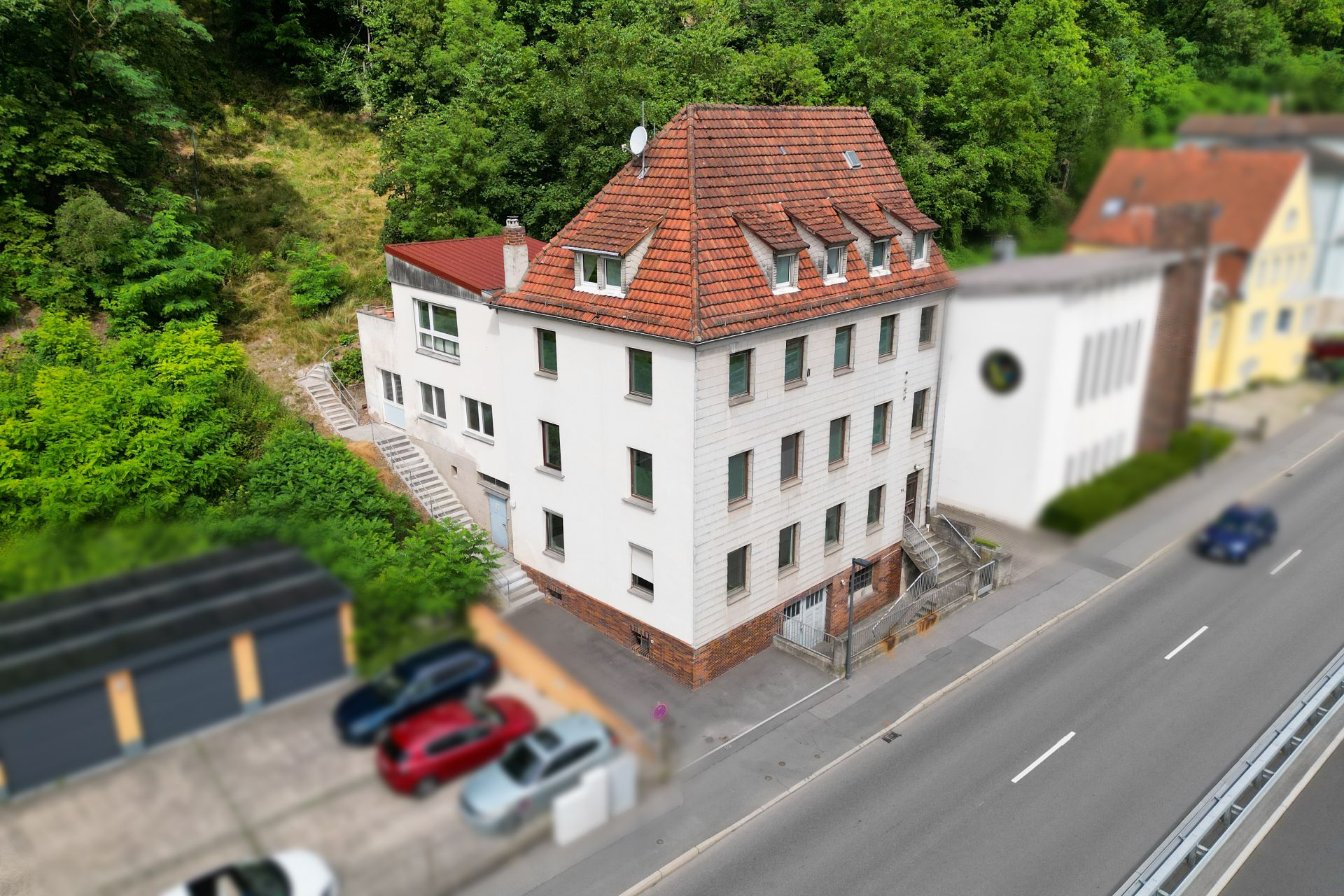 Mehrstöckiges Wohnhaus mit rotem Dach an einer Straße, umgeben von Bäumen und Nachbarhäusern.