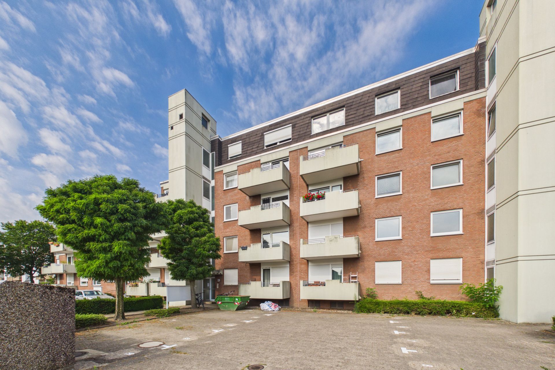 Mehrfamilienhaus mit Balkonen und Bäumen im Vordergrund, blauer Himmel darüber.