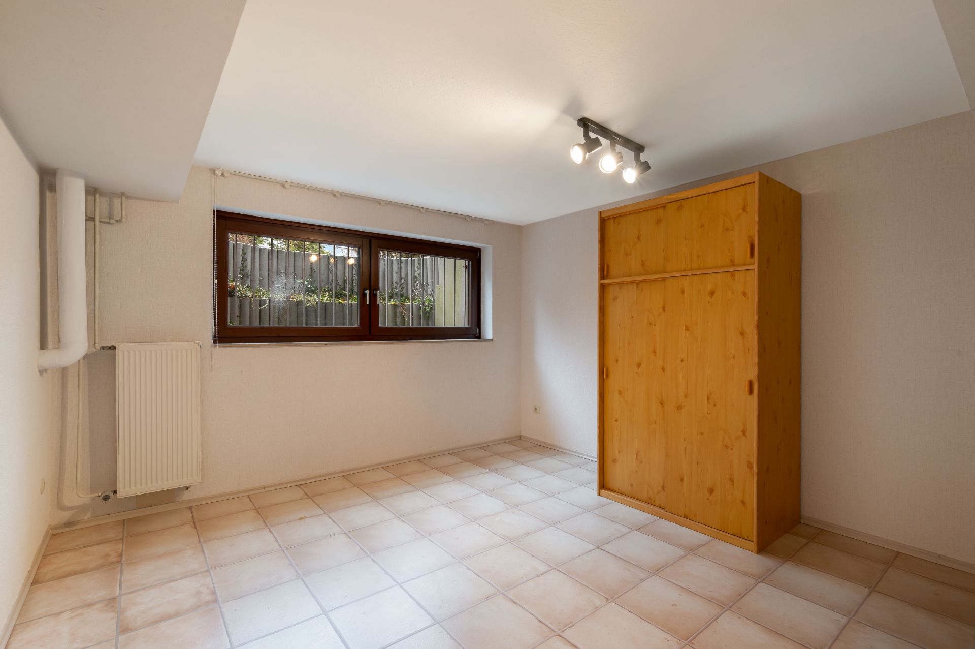 Helles Zimmer mit Fliesenboden, Holzschrank, großem Fenster und Deckenbeleuchtung.