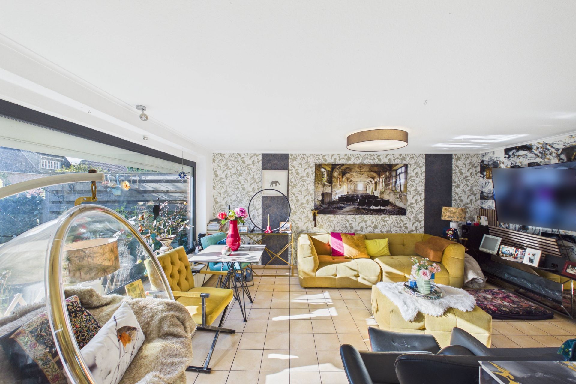 Helles Wohnzimmer mit gelbem Sofa, Glaskugelstuhl, großem Fenster und Wanddeko.