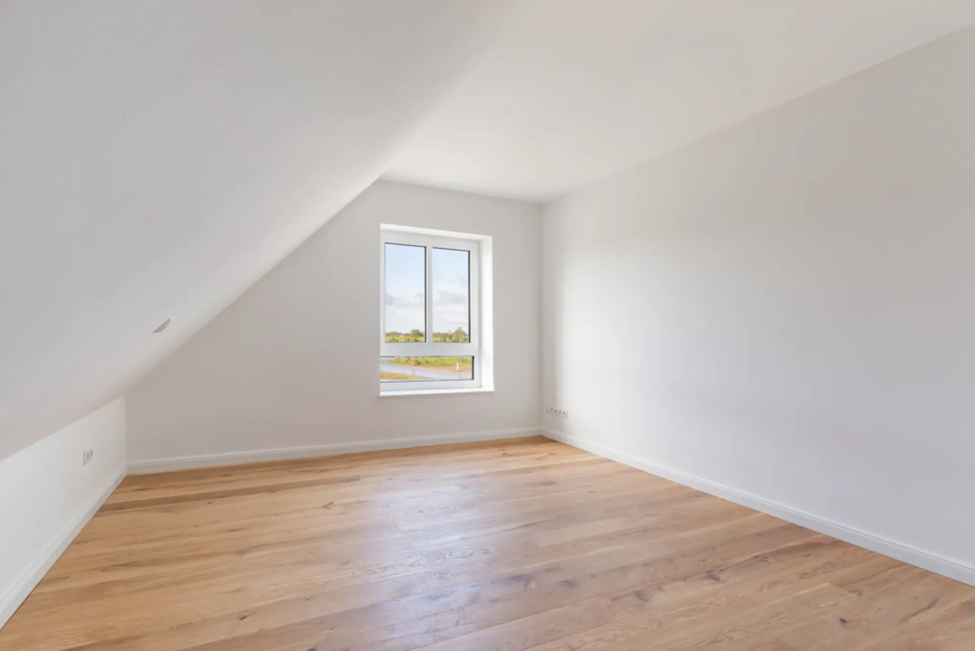 Leeres Zimmer mit schräger Decke, Holzfußboden und Fenster mit Blick ins Grüne.