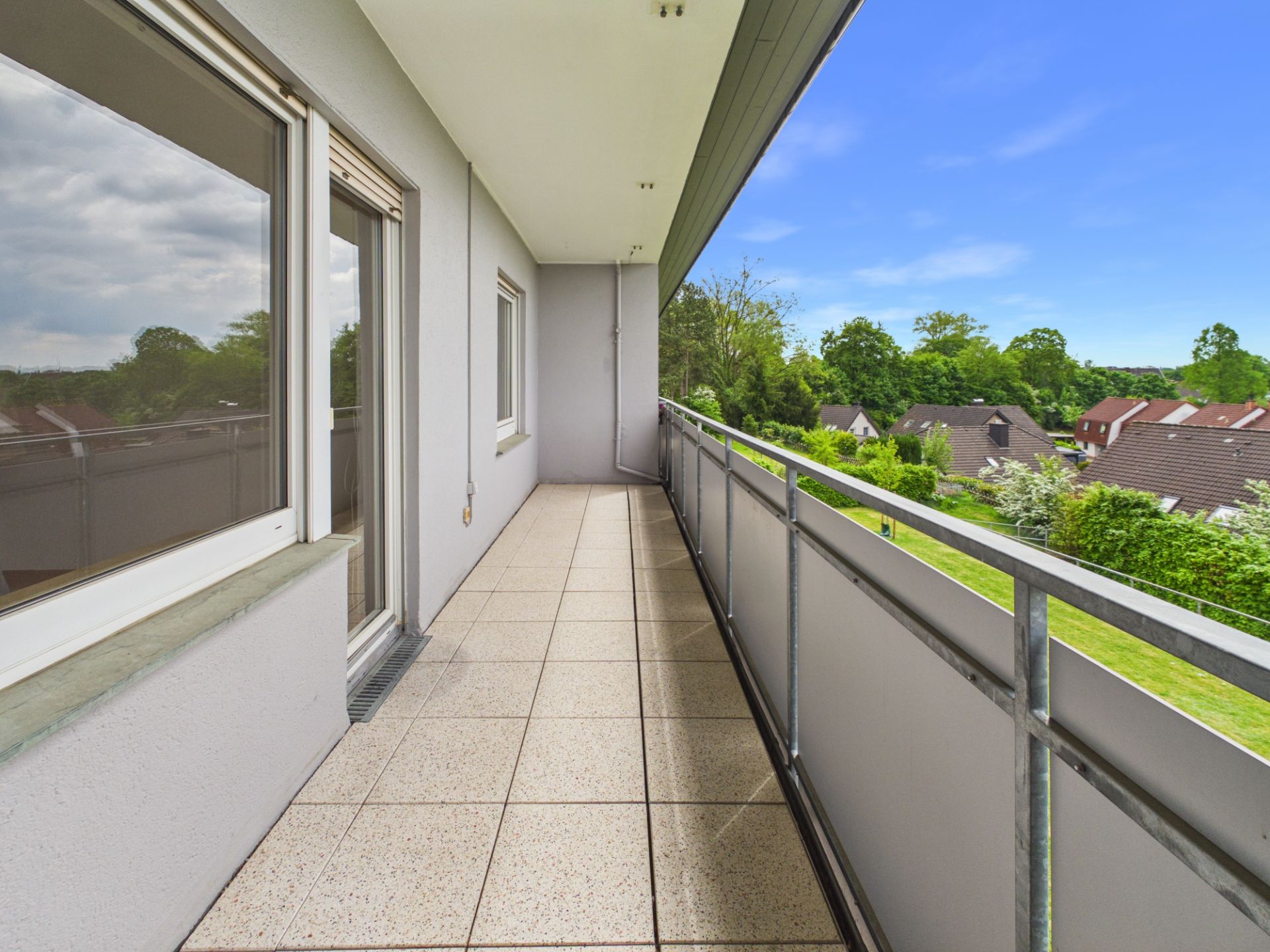 Langer Balkon mit Fliesenboden und Metallgeländer, Blick auf grüne Gärten und Nachbarhäuser.