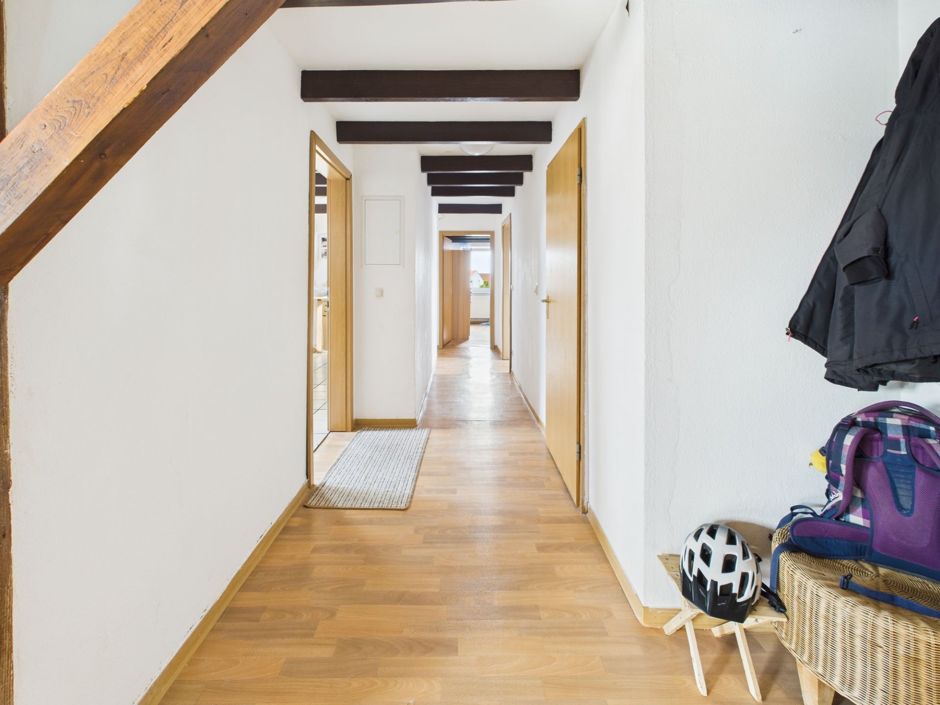 Langer Flur mit Holzboden, sichtbaren Deckenbalken, Türen und einer Garderobe mit Helm und Rucksack.