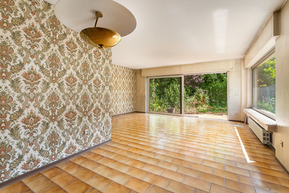 Leeres Wohnzimmer mit gemusterter Tapete, gelben Fliesen und großen Fenstern zum Garten.
