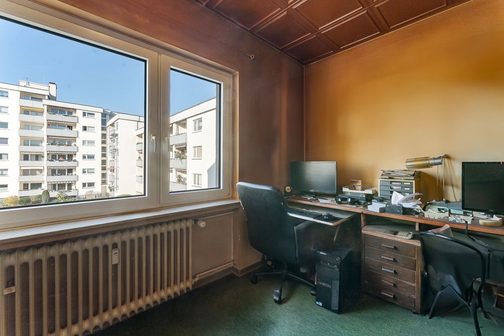 Büro mit Schreibtisch, zwei Monitoren und Blick aus dem Fenster auf Wohngebäude.