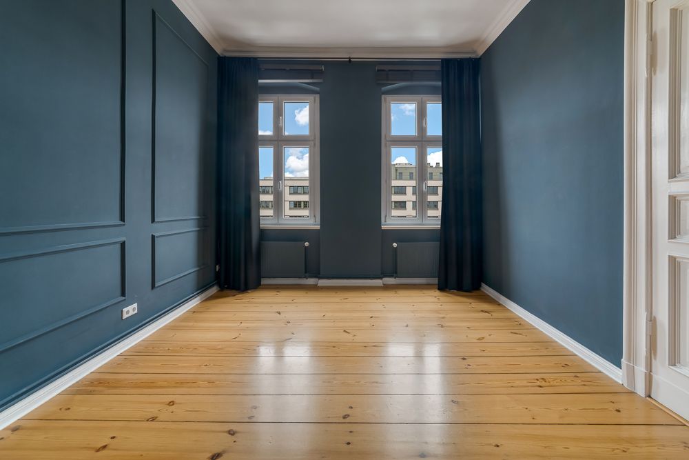 Leerer Raum mit Holzboden, blauen Wänden und zwei Fenstern mit dunklen Vorhängen.
