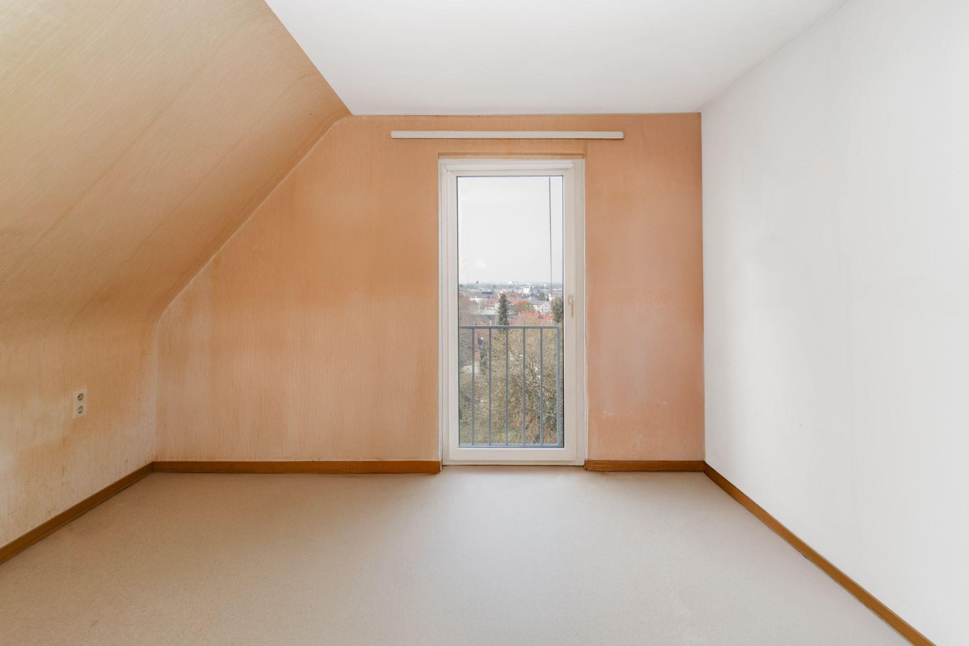 Leeres Zimmer mit Dachschräge und bodentiefem Fenster mit Blick nach draußen.