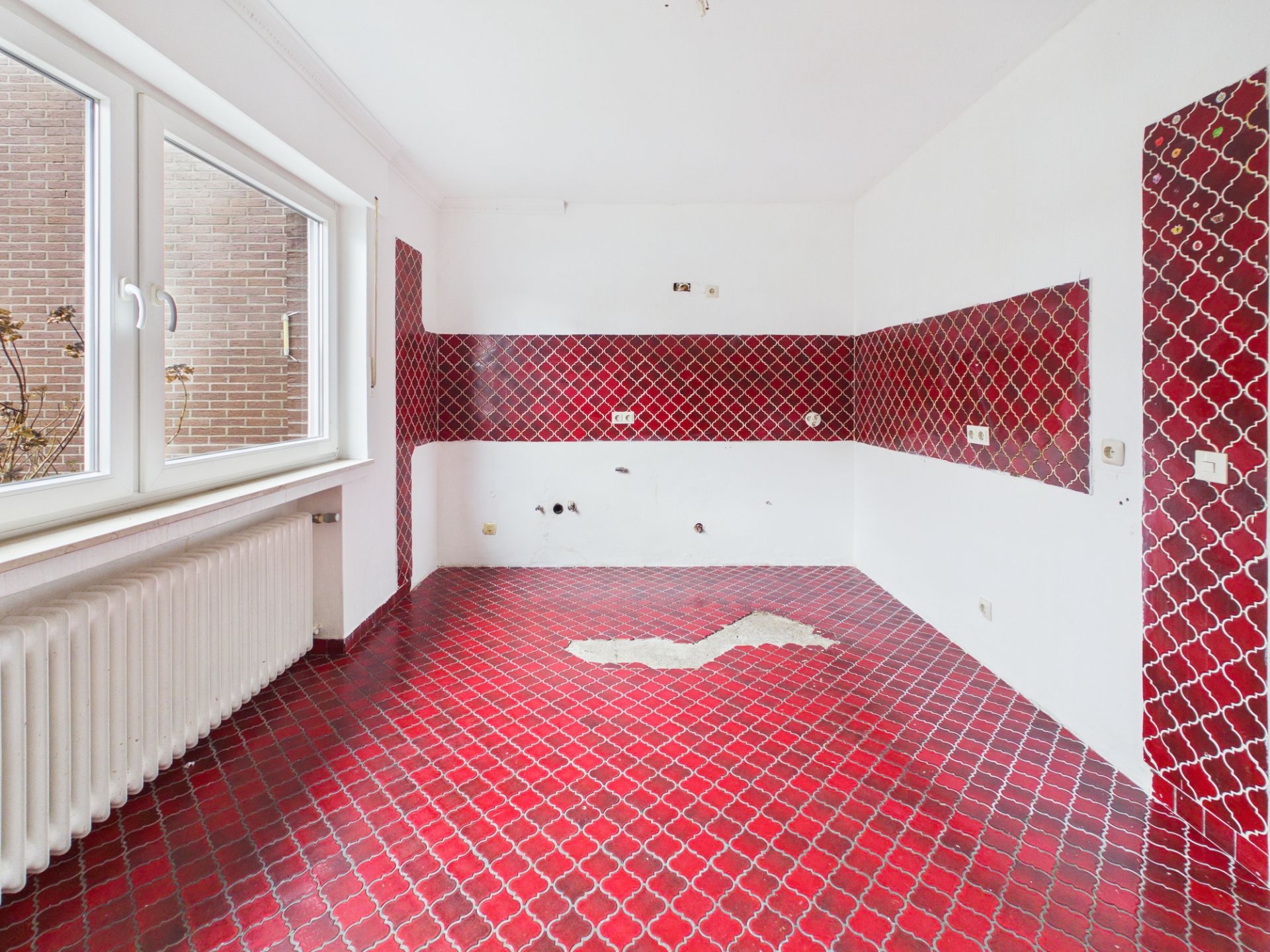 Leerer Raum mit roten Fliesen an Boden und Wänden, große Fenster und Heizkörper.
