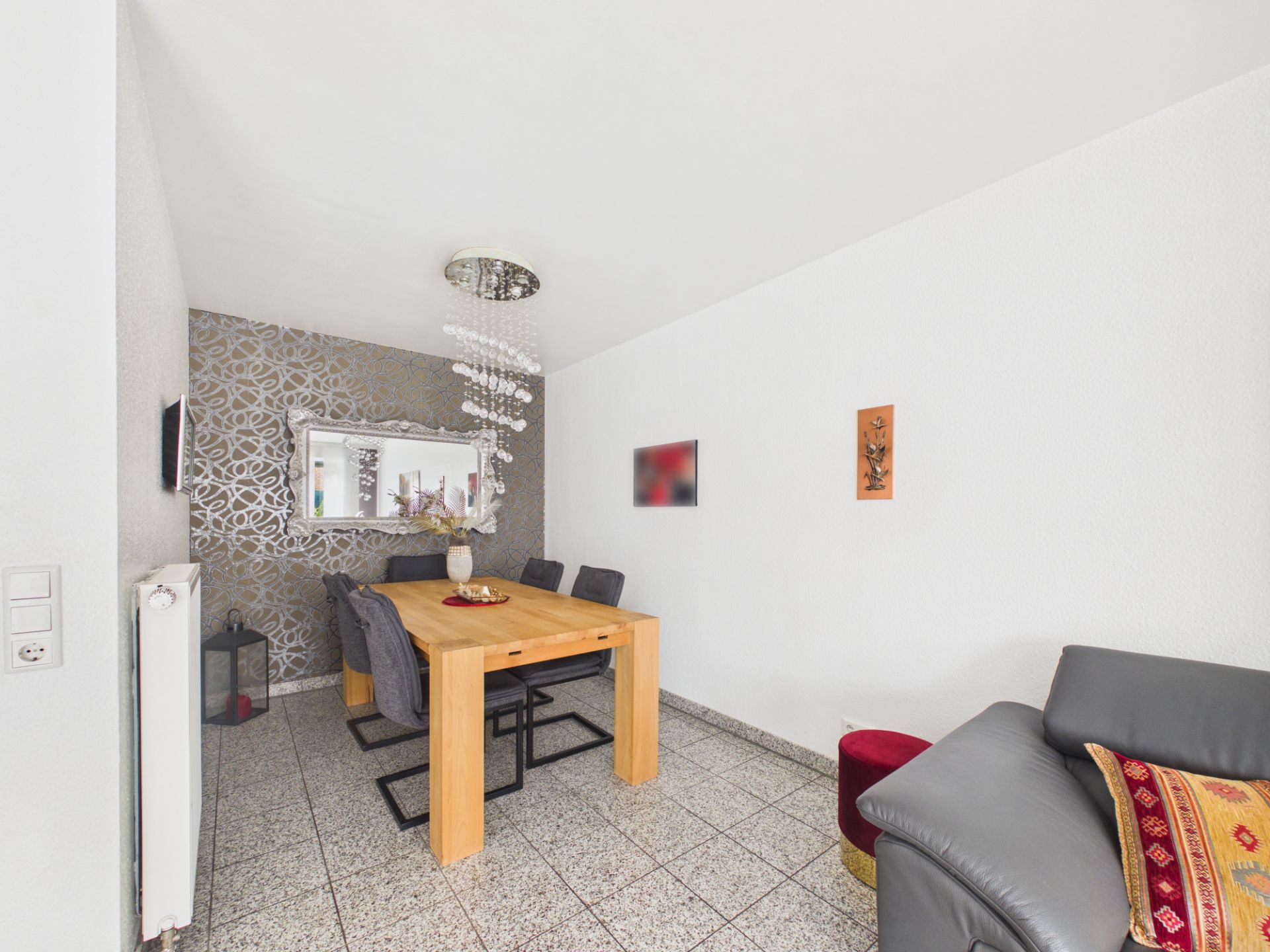 Essbereich mit Holztisch, sechs Stühlen, großer Spiegel und grauem Sofa auf Fliesenboden.
