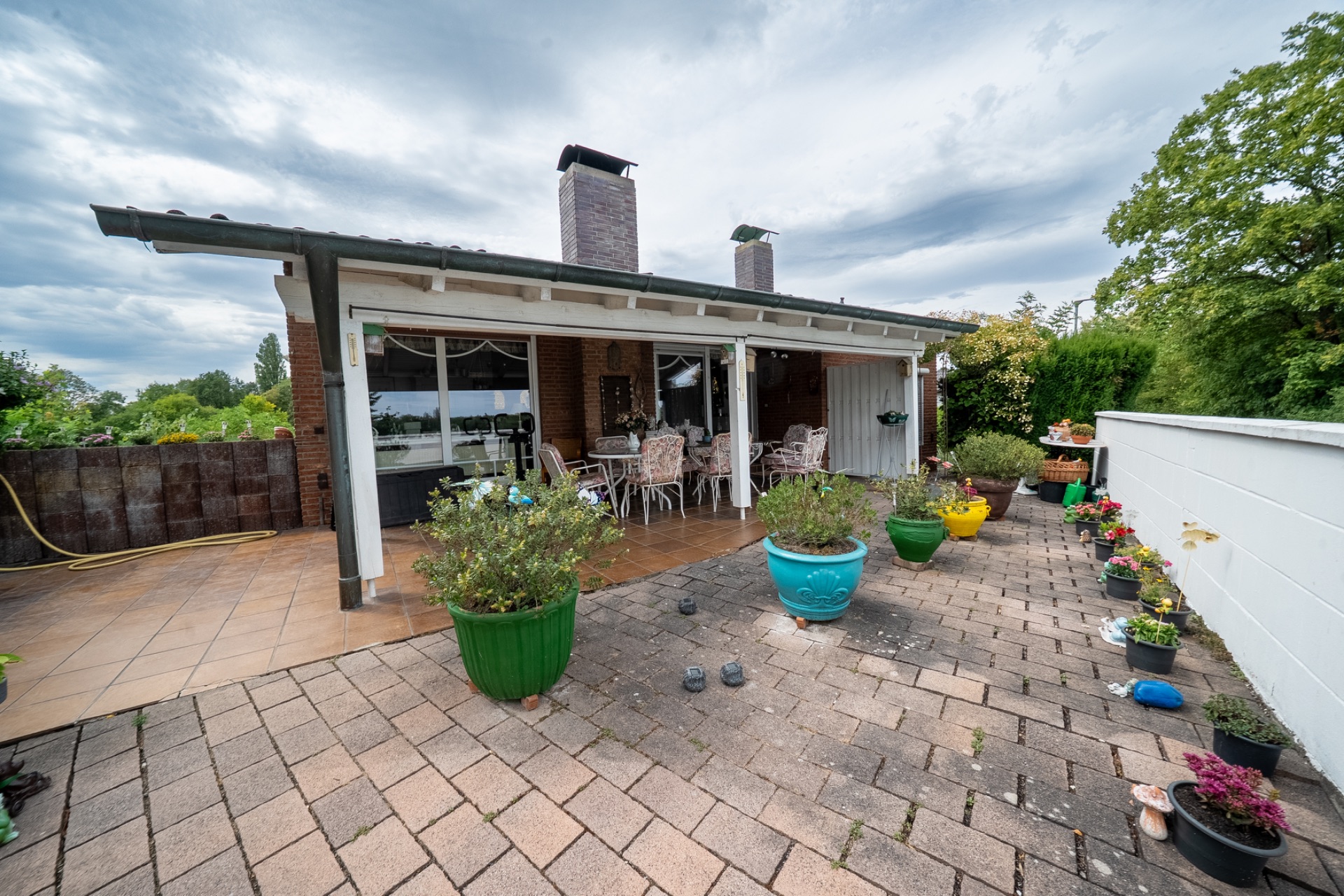 Überdachte Terrasse mit Gartenmöbeln und vielen bunten Blumentöpfen auf gepflastertem Boden.