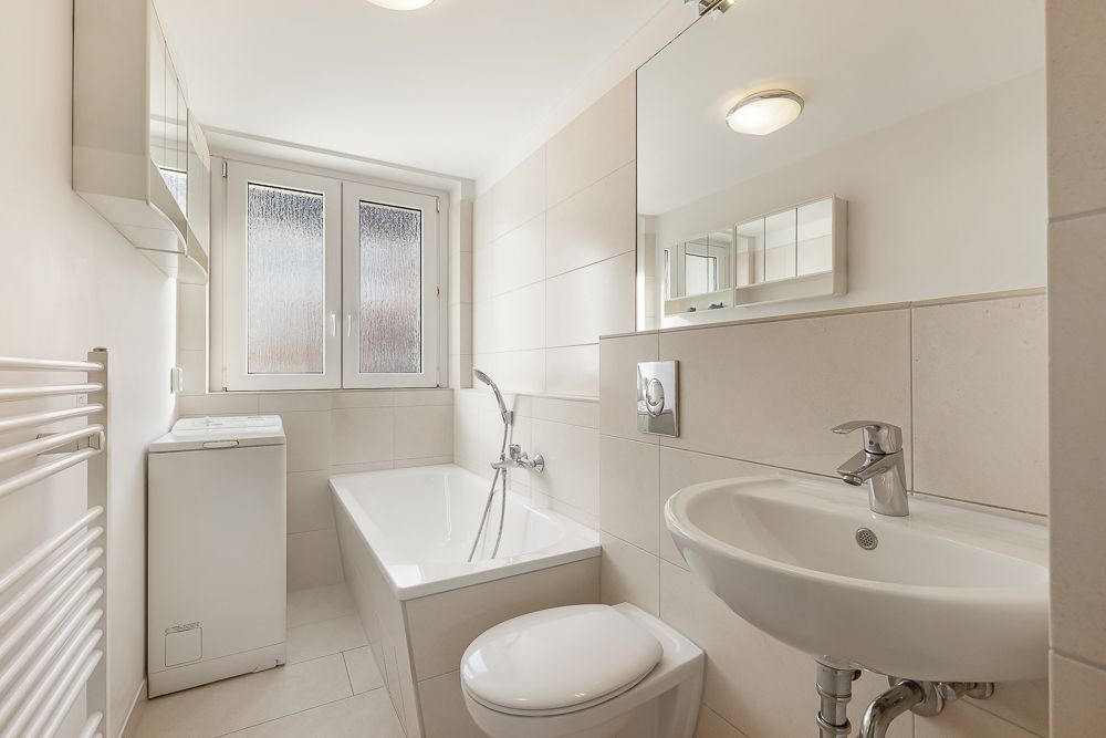 Helles Badezimmer mit Badewanne, Waschmaschine, Toilette, Waschbecken und Fenster.