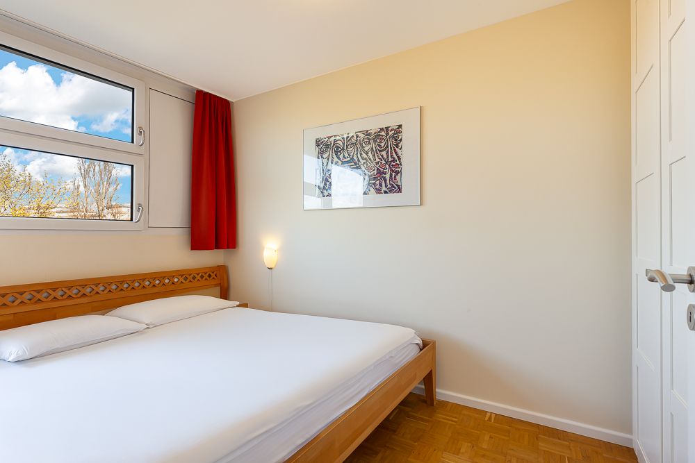 Helles Schlafzimmer mit Doppelbett, Fenster, rotem Vorhang und Wandbild.