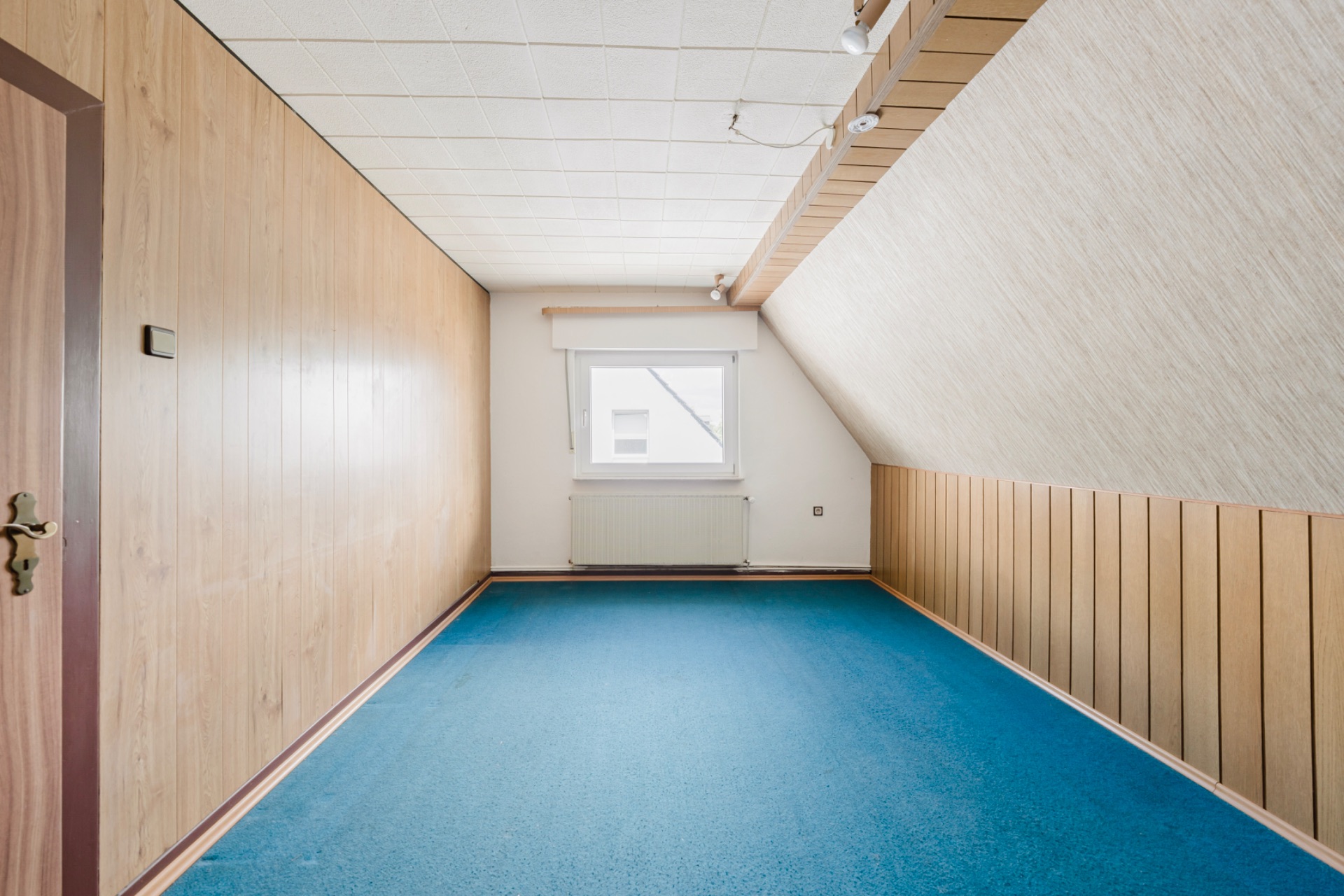 Leerer Raum mit blauer Teppich, Holzvertäfelung und kleinem Fenster an der Stirnwand.