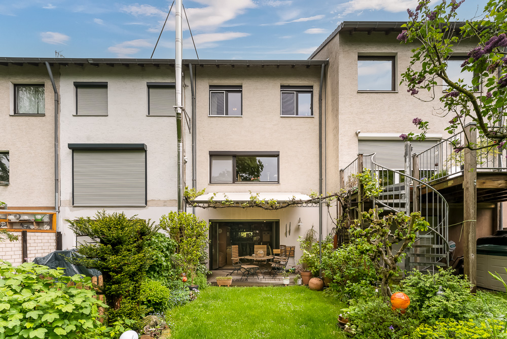 Rückseite eines Hauses mit Garten, Terrasse und Gartenmöbeln, umgeben von Pflanzen.