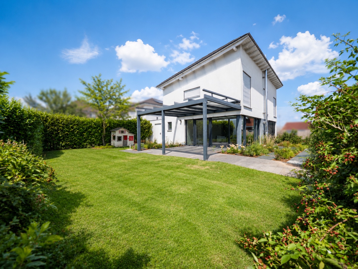 Modernes weißes Wohnhaus mit Garten, Terrasse und Kinderspielhaus bei sonnigem Wetter.