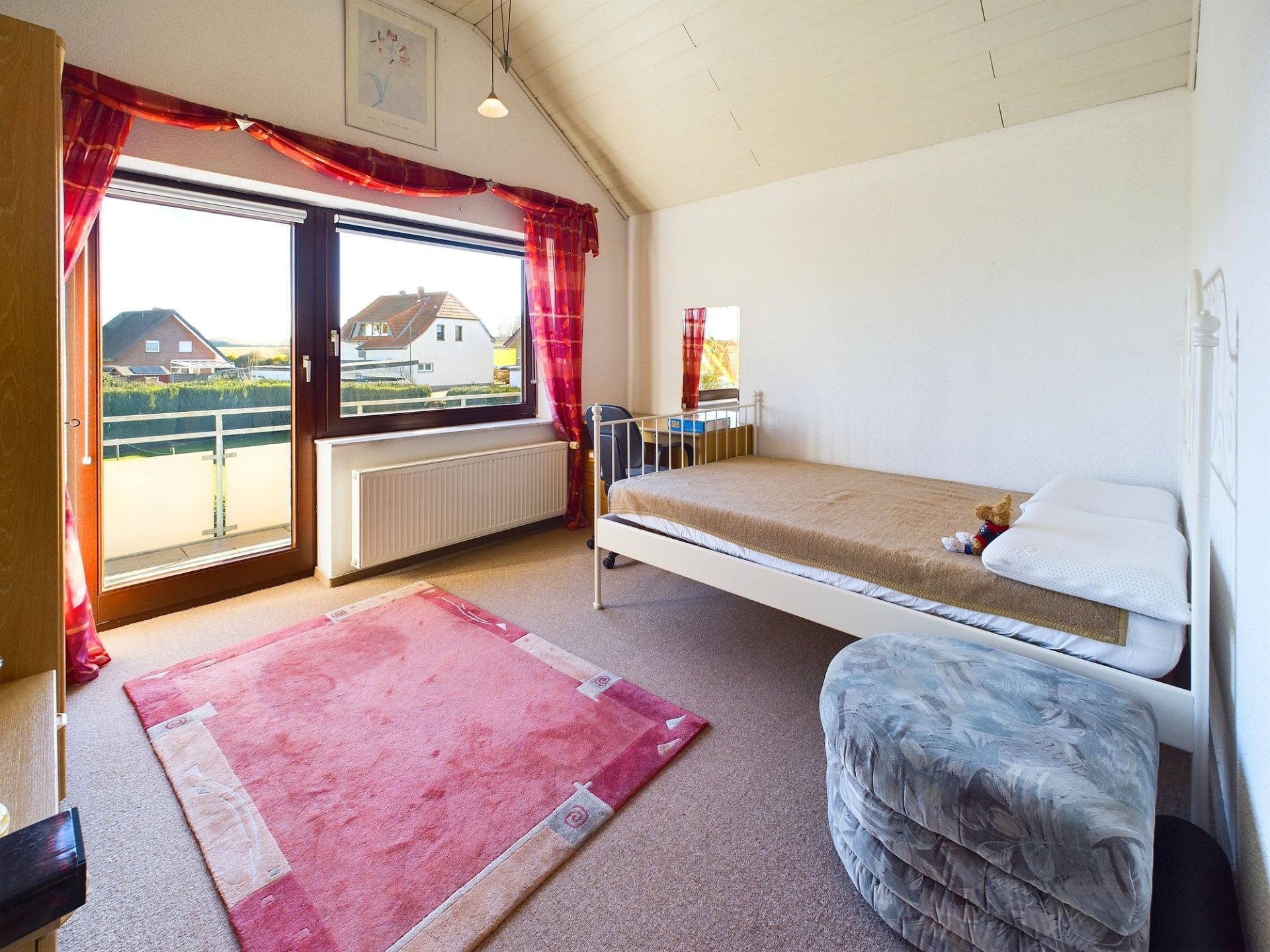 Helles Schlafzimmer mit Einzelbett, rotem Teppich, Hocker und großem Fenster mit Blick nach draußen.