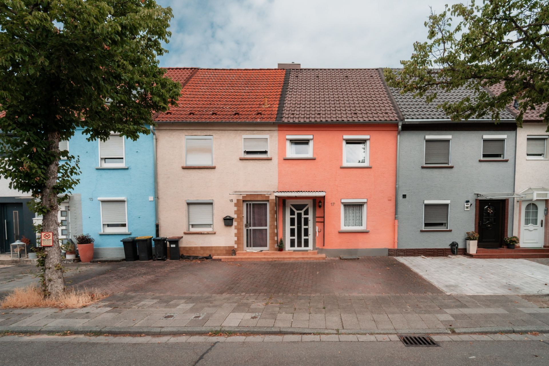 Reihenhäuser in verschiedenen Farben mit gepflasterter Einfahrt und Bäumen am Straßenrand.