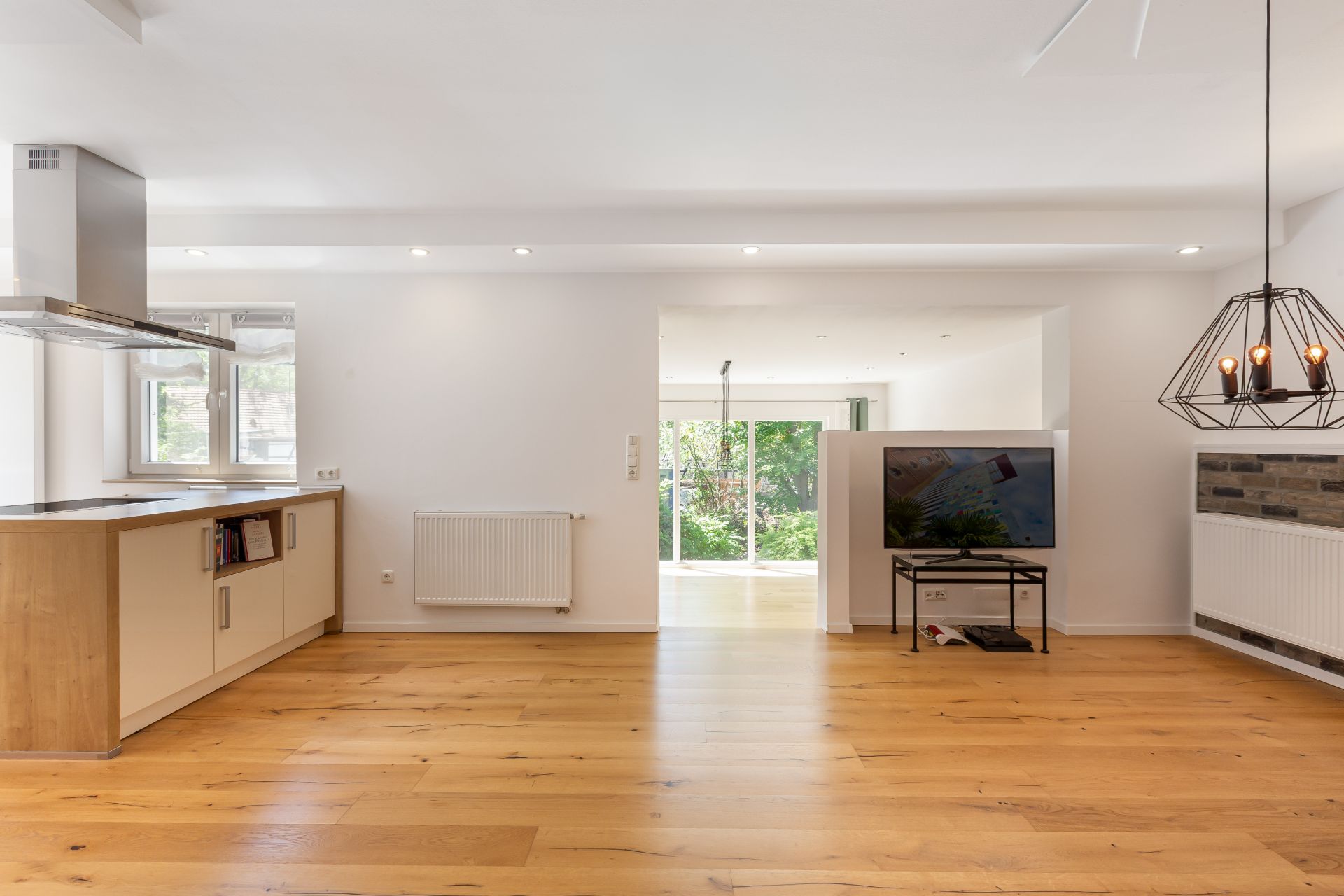 Helles Wohnzimmer mit offenem Küchenbereich, Holzfußboden, Fernseher und großen Fenstern.