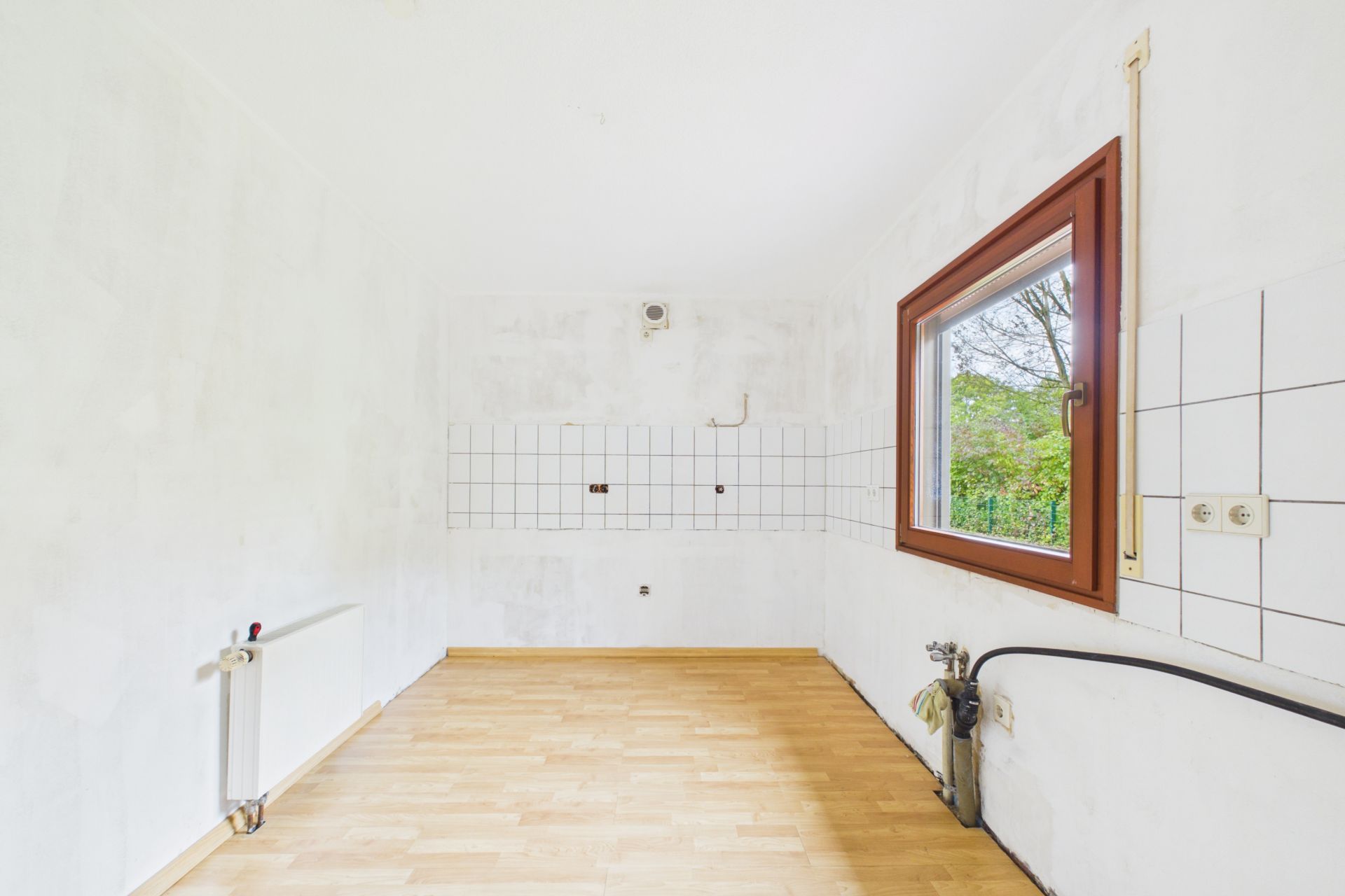 Leerer Raum mit Fliesen an der Wand, Heizkörper und Fenster mit Blick ins Grüne.