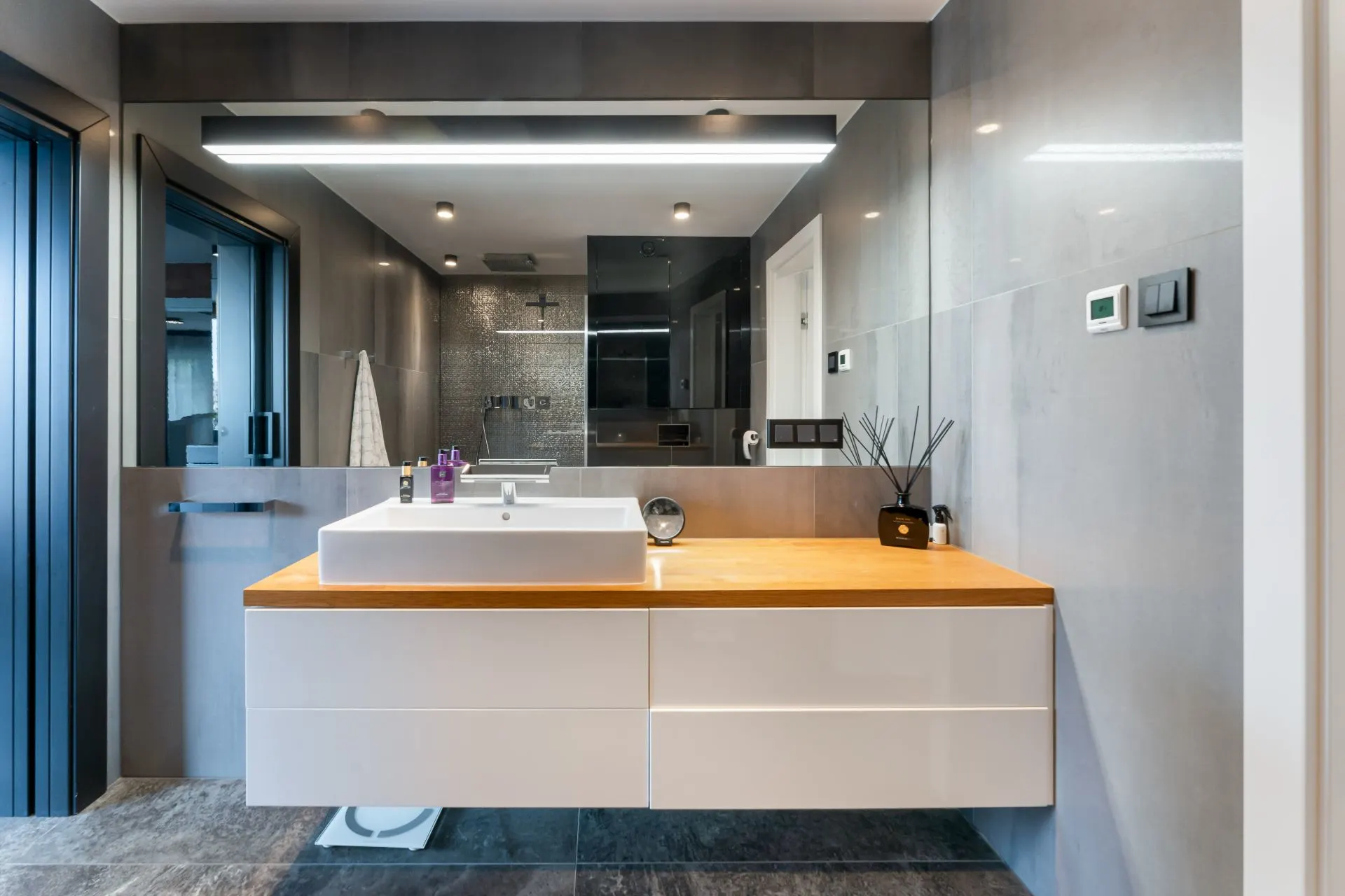 Modernes Badezimmer mit großem Spiegel, Waschbecken auf Holzplatte und grauen Fliesen.