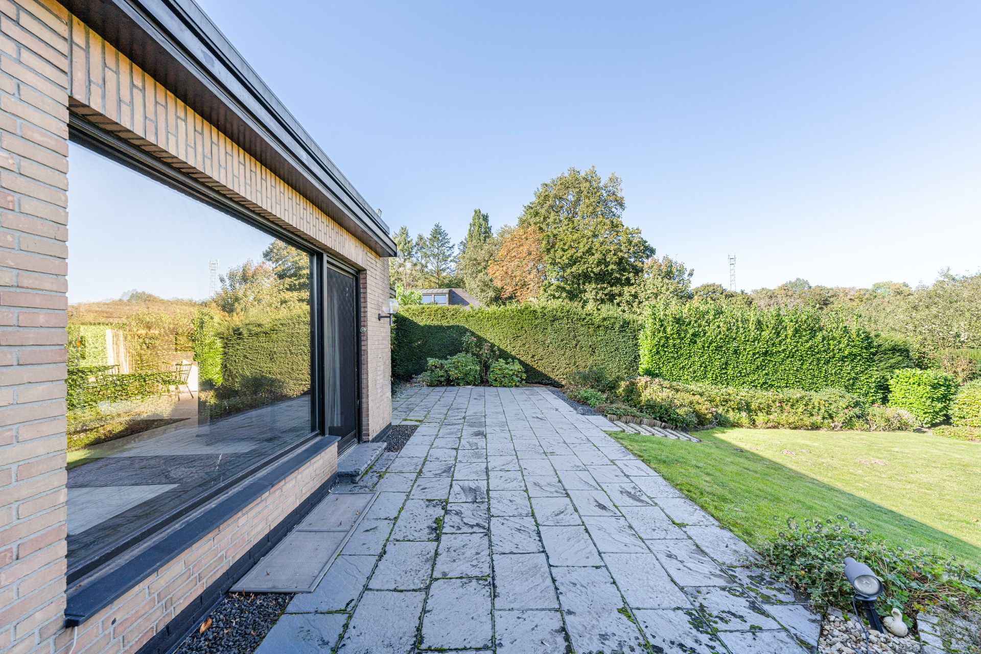 Gepflasterte Terrasse neben einem Haus mit Garten und grüner Hecke im Hintergrund.