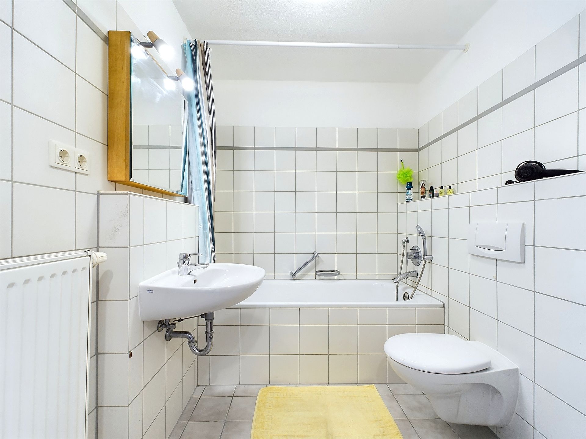 Helles Badezimmer mit Badewanne, Wandspiegel, Waschbecken und Toilette, gelber Teppich am Boden.