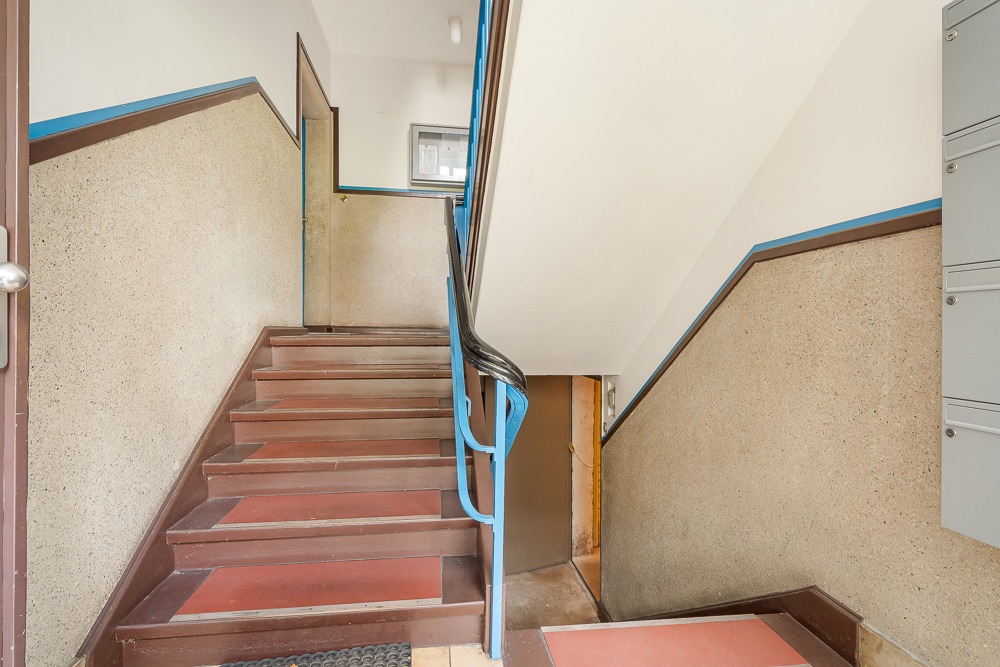 Treppenhaus eines Wohngebäudes mit braunen Stufen und blauem Handlauf.